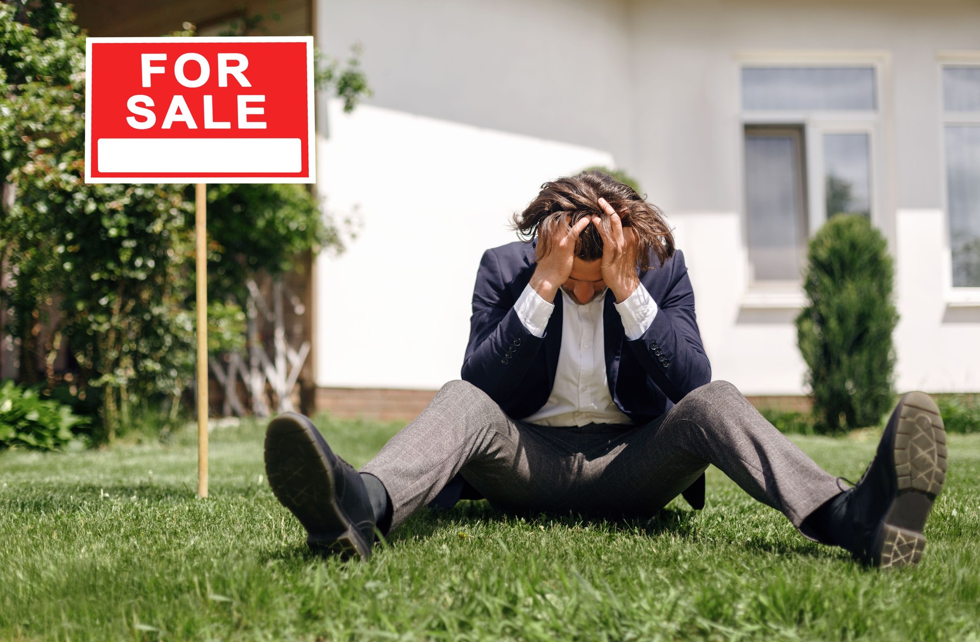 Depressed real estate agent sitting on grass in front of house for sale, outdoors